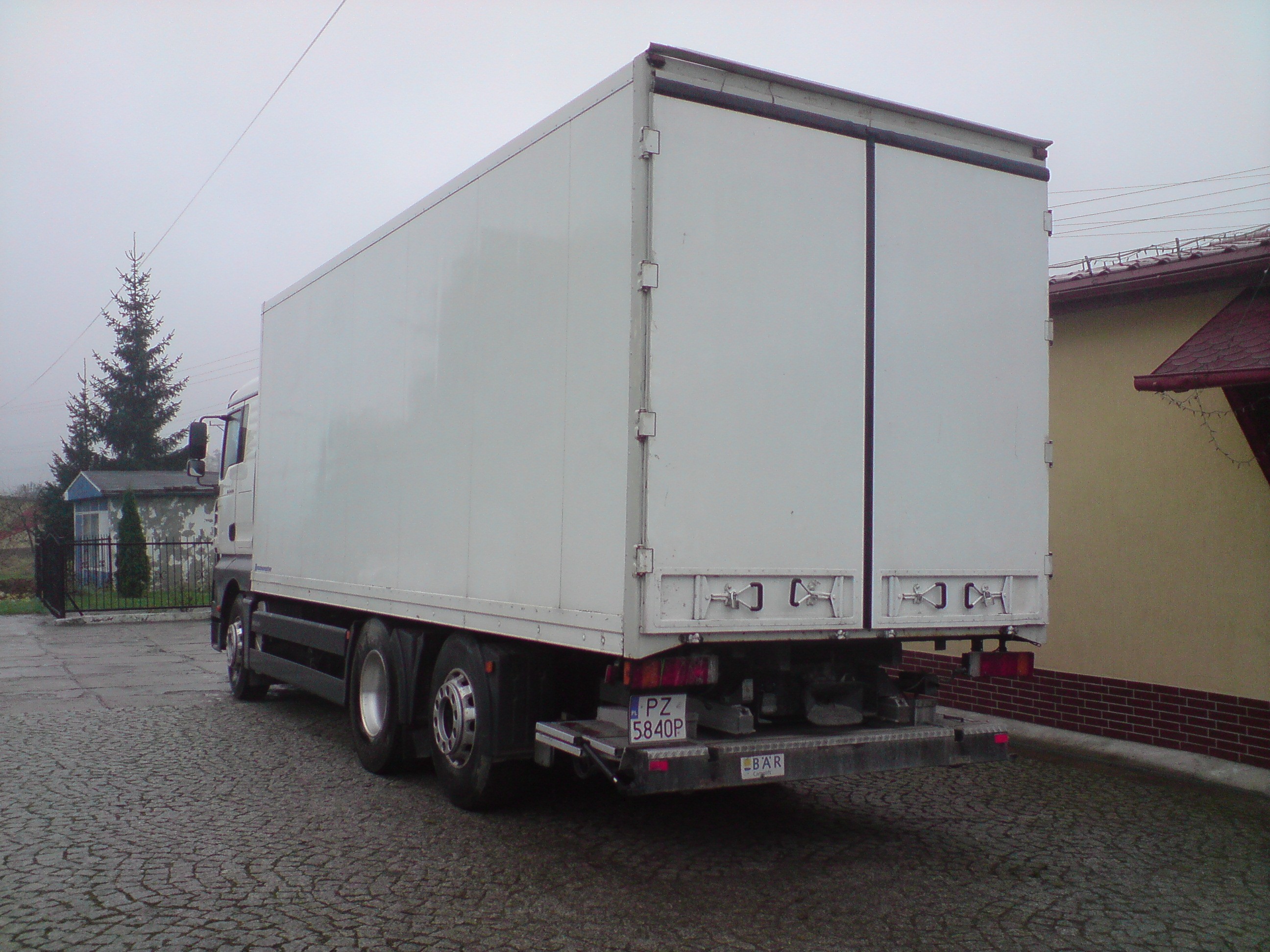 Tył białej ciężarówki z zabudową kontenerową, zaparkowanej na brukowanej nawierzchni w pochmurny dzień, z widocznym numerem rejestracyjnym i napisem 'BAR' na tylnym zderzaku, na tle budynku...